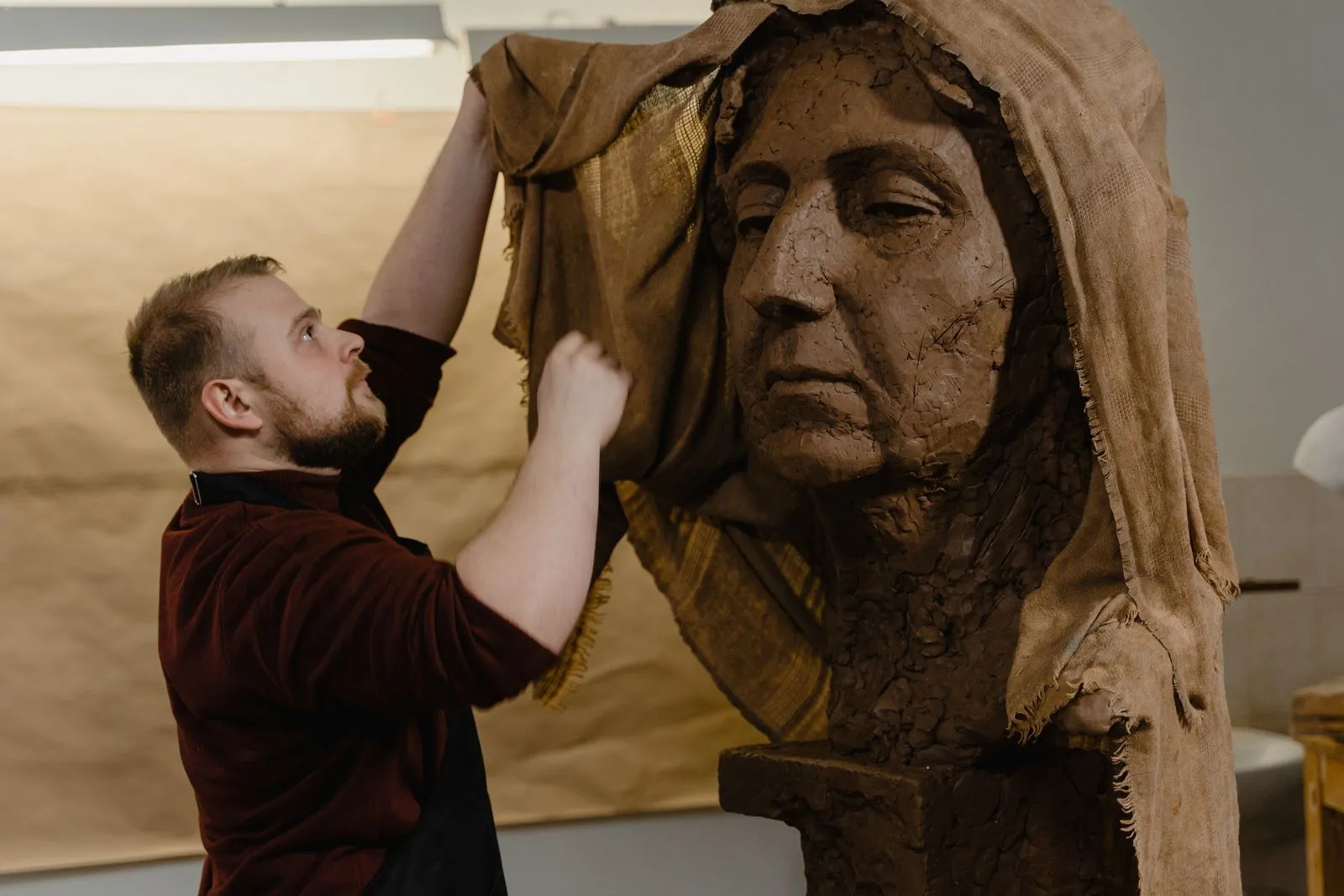 A sculptor carefully unveils a clay bust in a studio, showcasing artistry and craftsmanship.